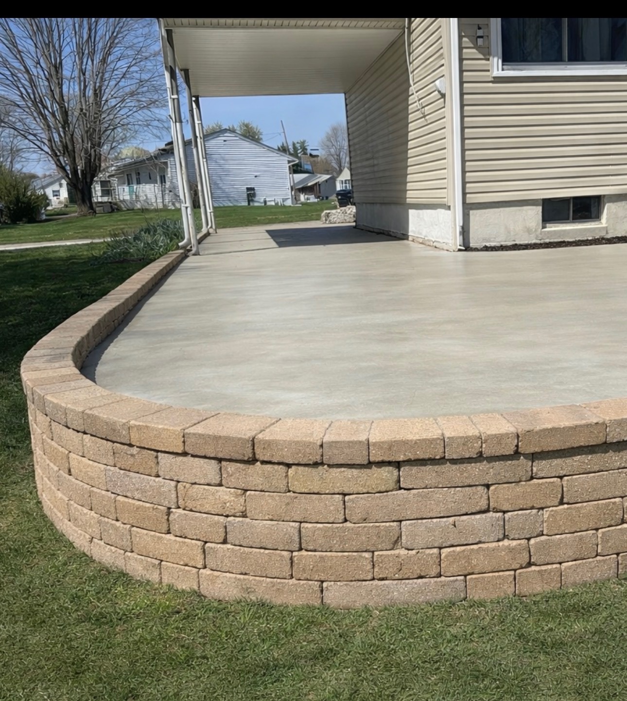 Curved stone retaining wall with a concrete patio beside a tan house and green lawn.