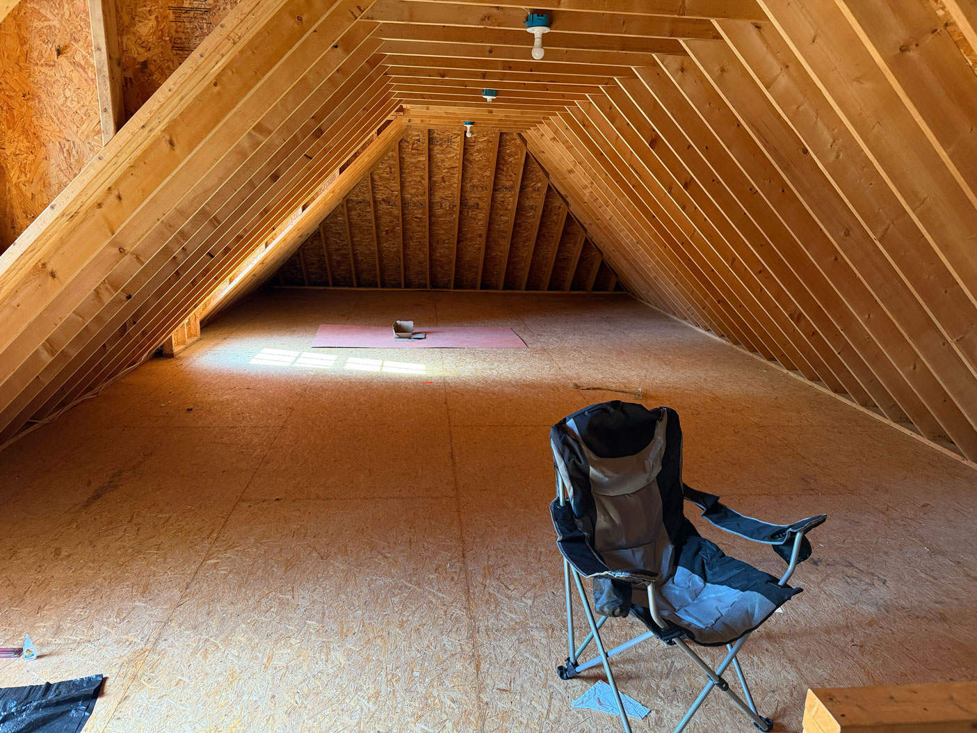 Unfinished attic with wooden beams, a folding chair, and sunlight coming through a window on the floor.