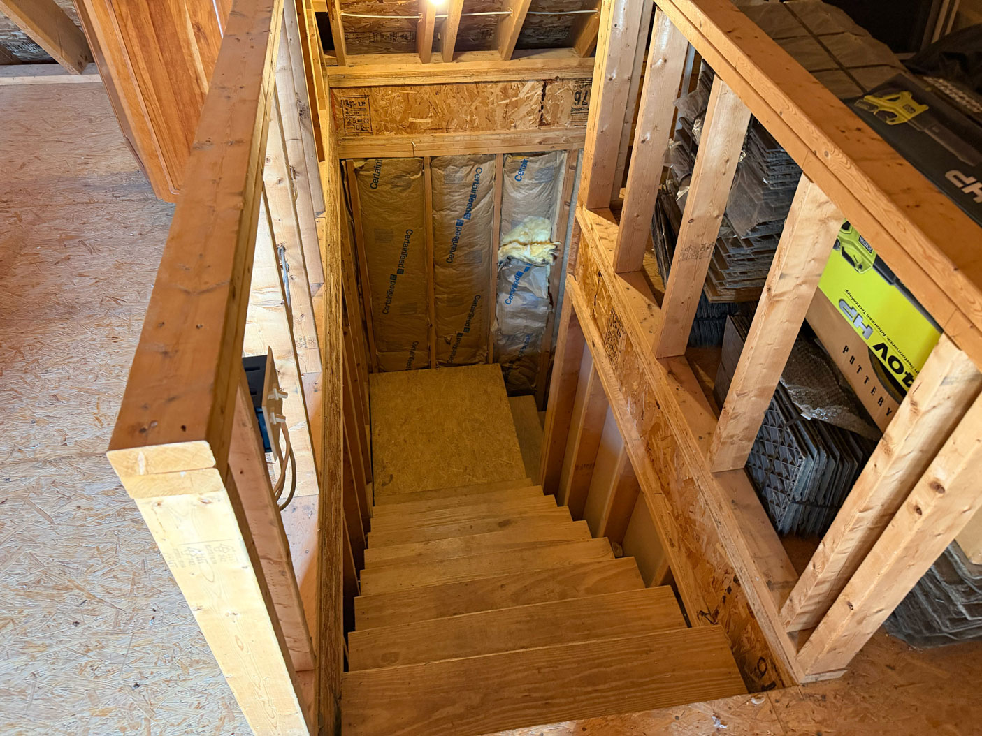 Wooden stairs leading down in an unfinished attic with exposed beams, insulation, and building materials nearby.