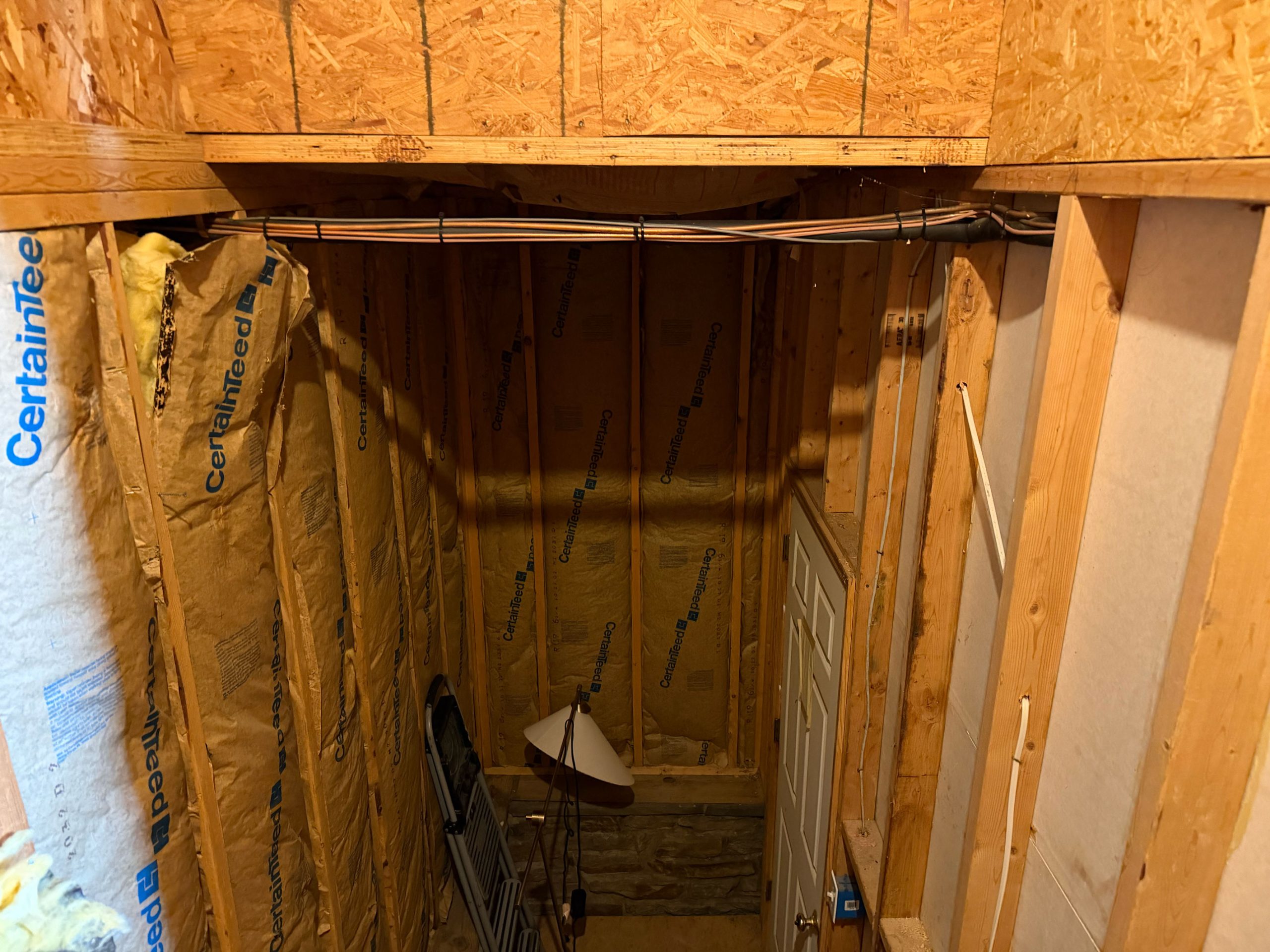 Unfinished basement with exposed insulation, wooden framing, a door, a floor lamp, and a folded metal chair.