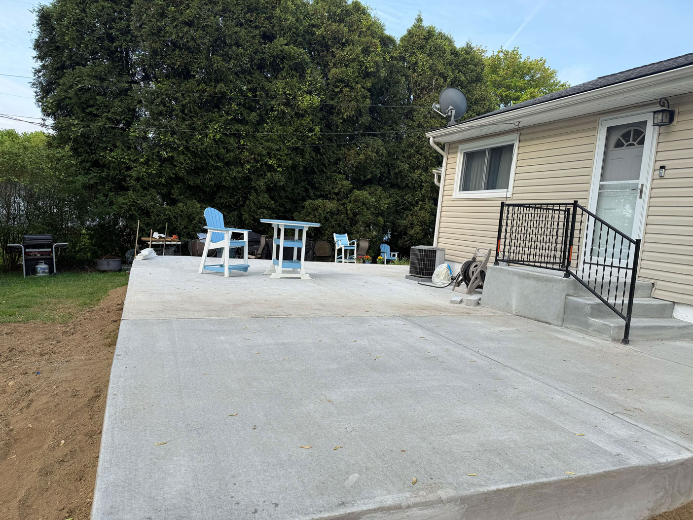 A concrete patio with blue chairs and a table beside a beige house, surrounded by green trees.