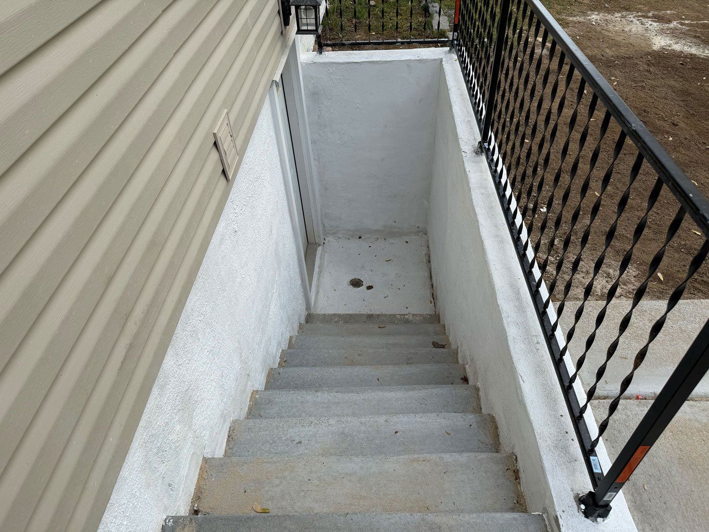 Concrete stairs lead down to a basement door, surrounded by white walls and a black metal railing.