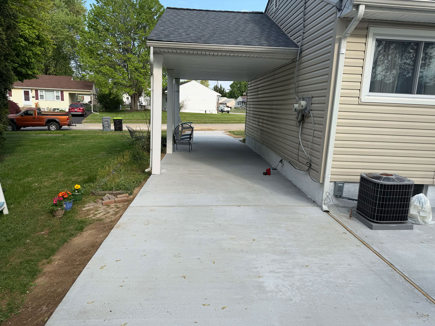 Concrete driveway beside a tan house with a covered carport, chair, flowers, and green lawn.