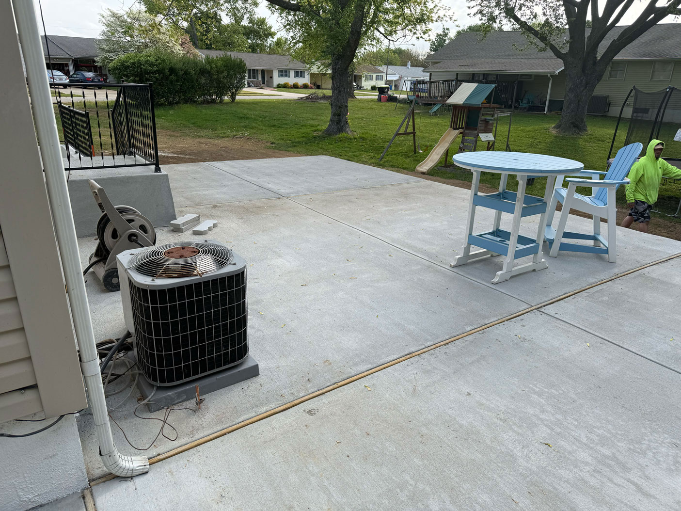 A concrete patio with a table, chairs, an AC unit, and a person in a green shirt near a playground.