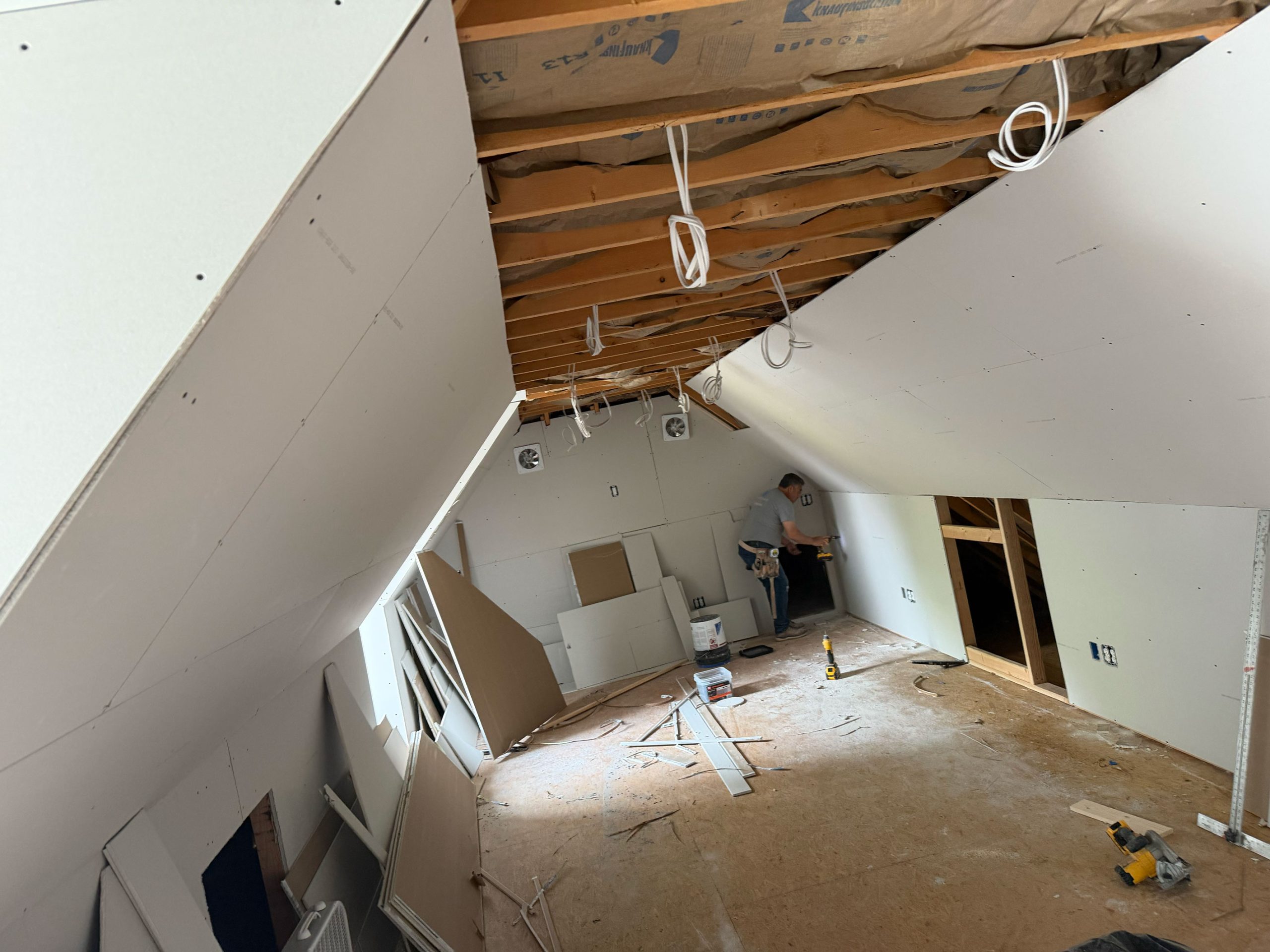 Person installing drywall on attic walls, with exposed insulation and construction materials scattered on the floor.