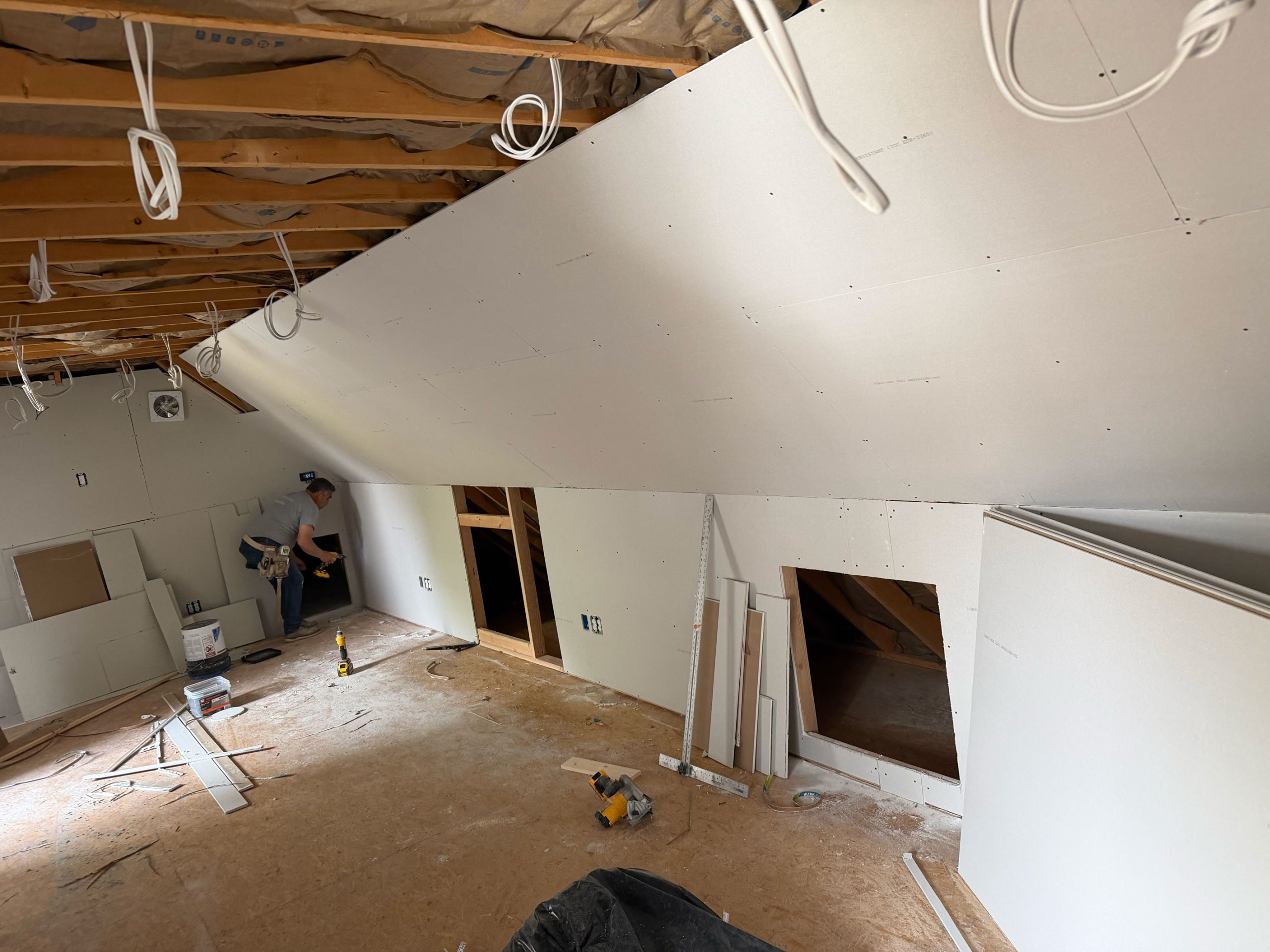 Person installing drywall on sloped ceiling in unfinished attic with exposed beams and construction tools.