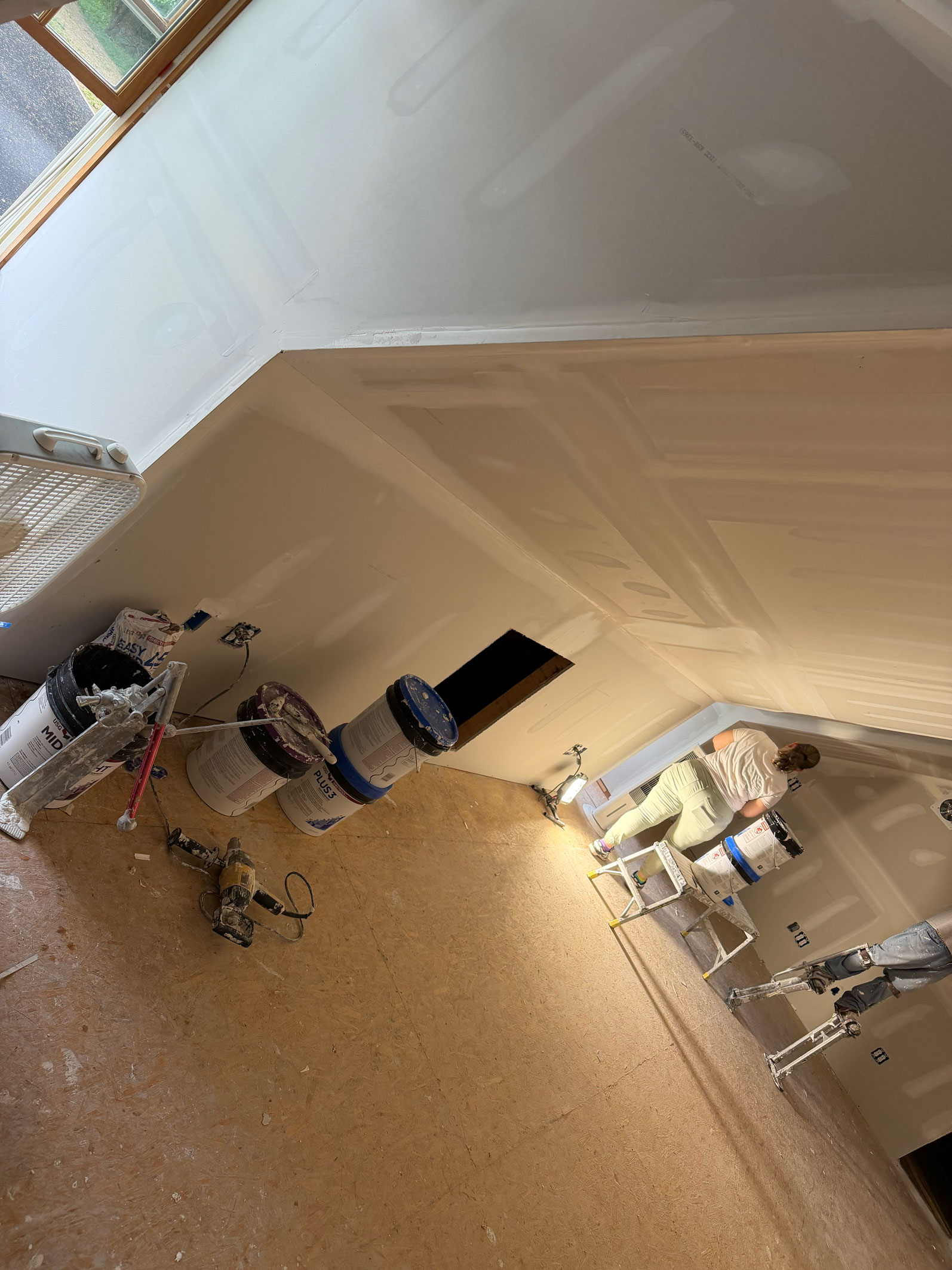 Person painting a room with drywall, paint cans, and equipment on the floor; sunlight streams in.