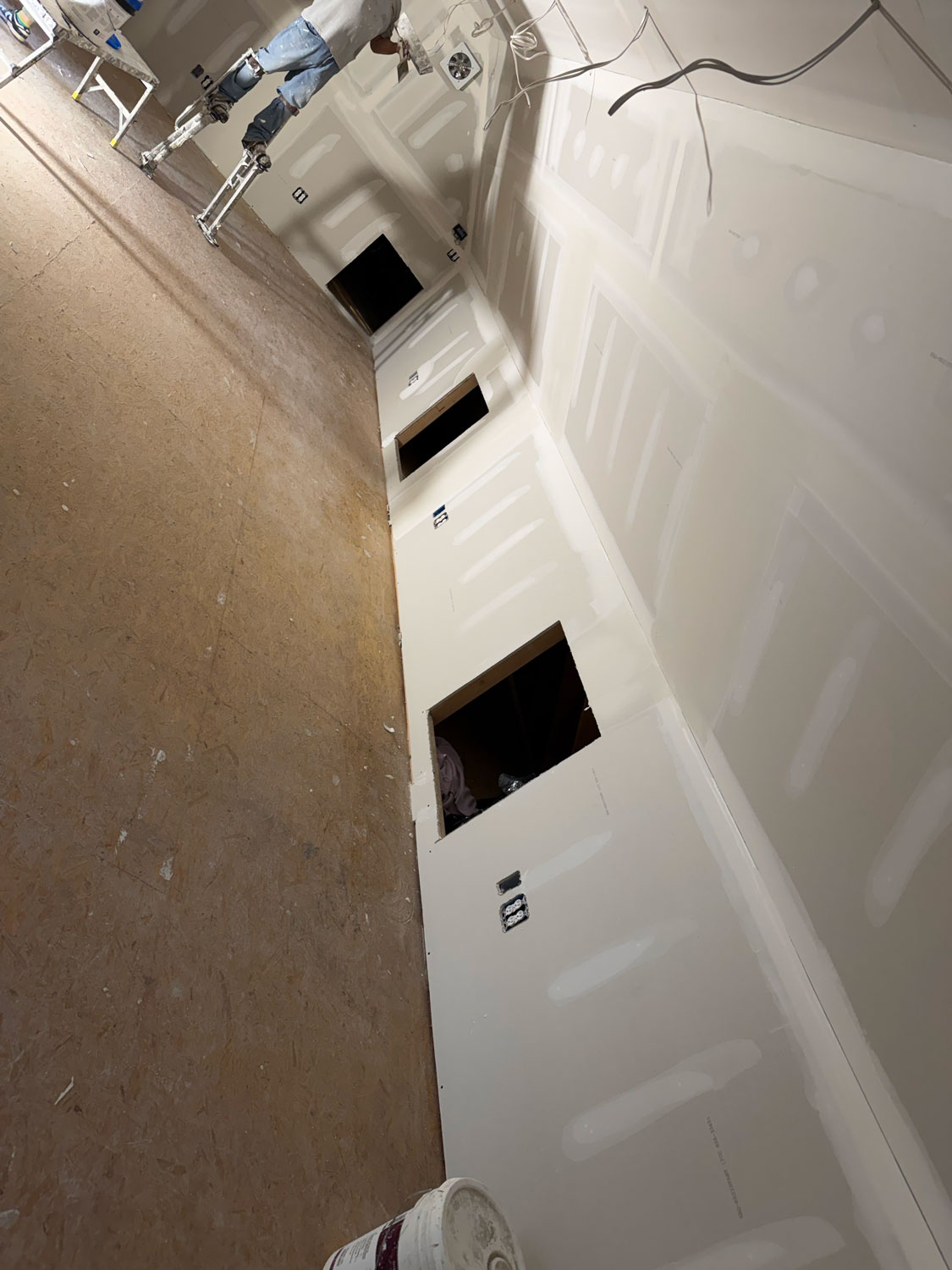 A room under construction with drywall, cutouts for outlets, and a person working on a ladder.