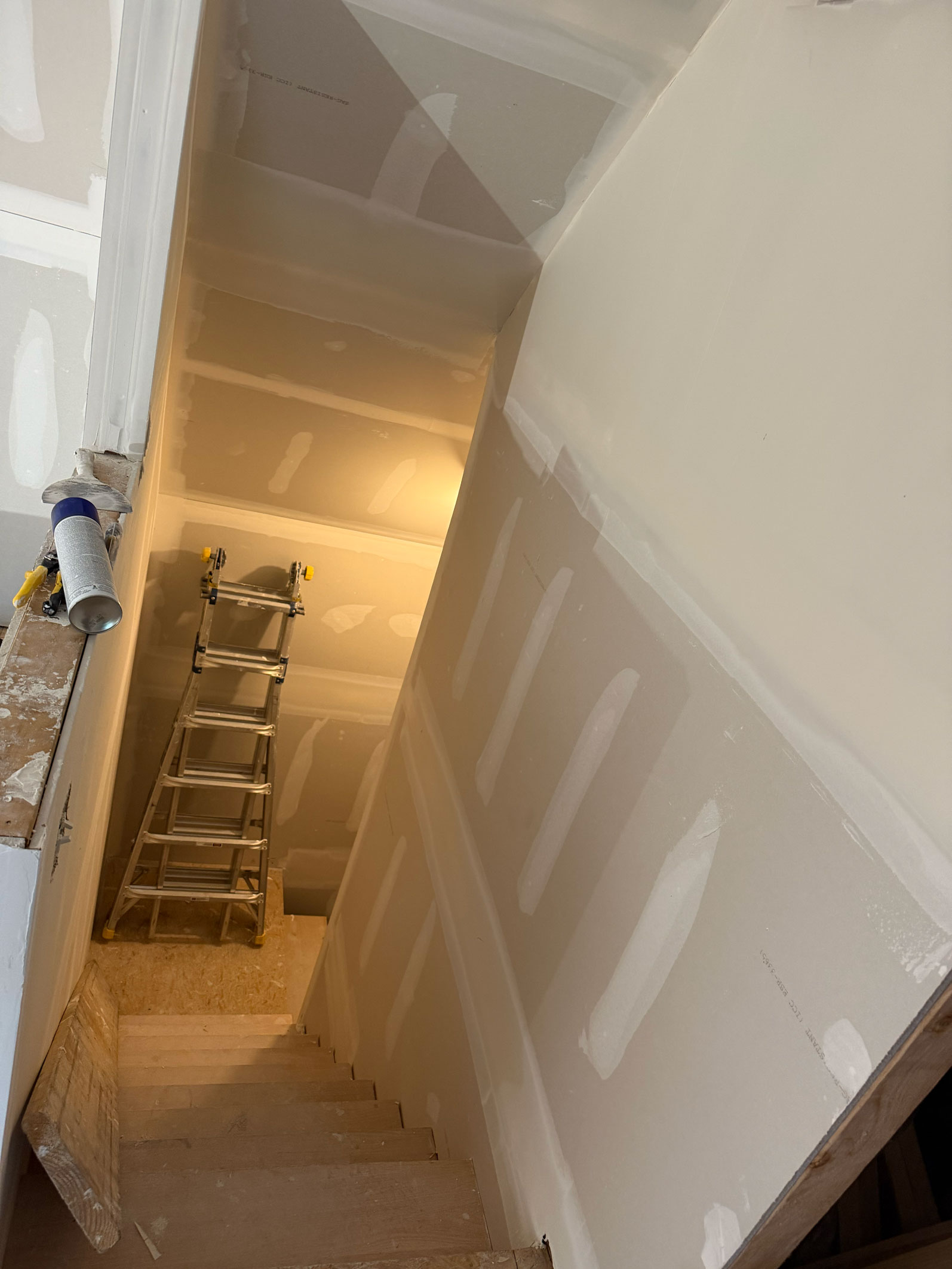 A stairwell under construction with drywall and a folded ladder at the bottom, lit by a single light.