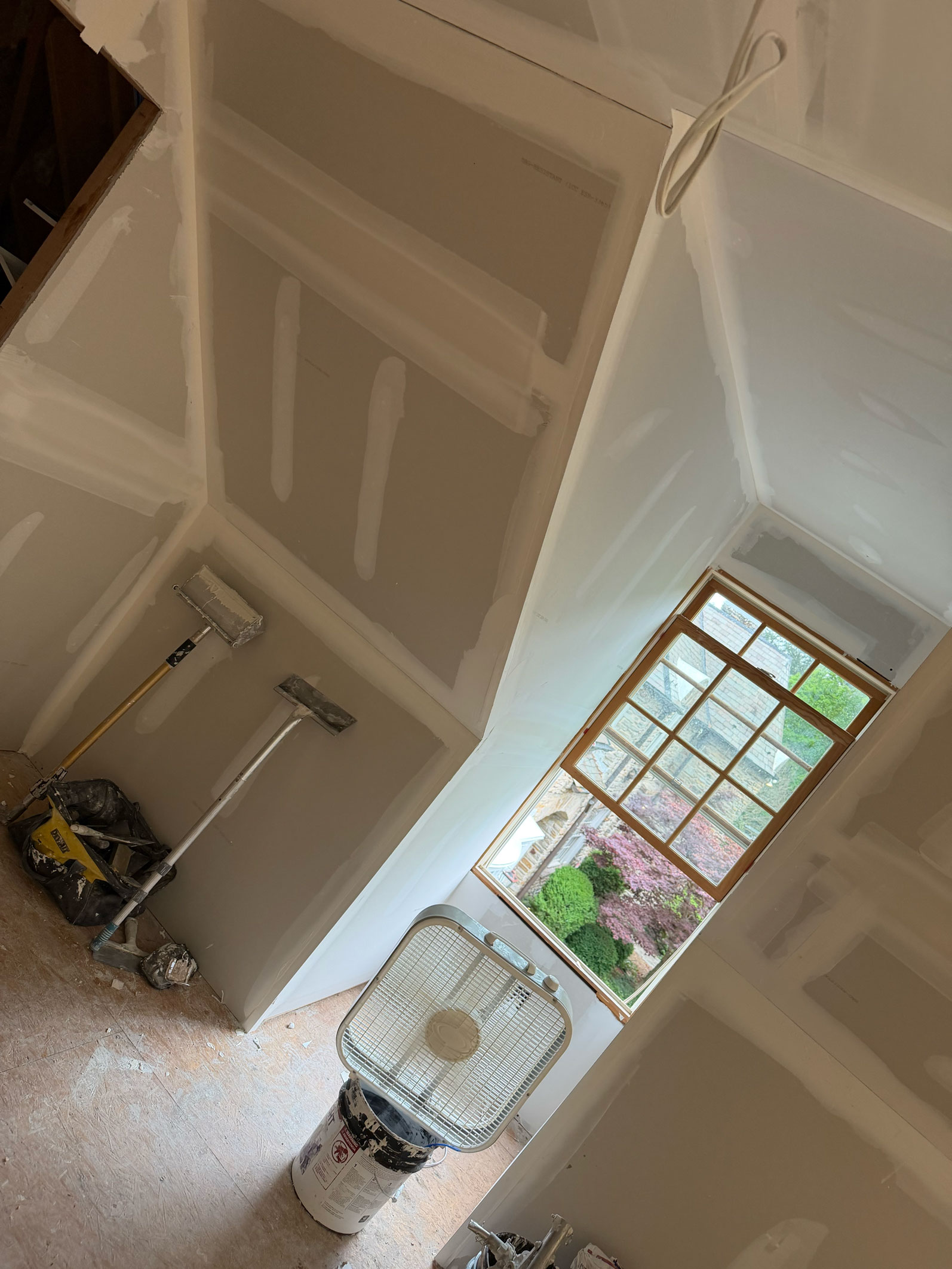 Room under construction with drywall, joint compound, painting tools, a fan, and a window showing greenery outside.