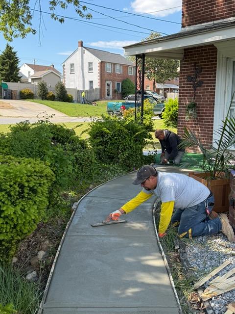 Two workers smooth wet concrete on a curved sidewalk in front of a brick house with green bushes nearby.