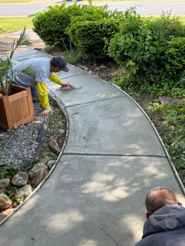 Two people smooth wet concrete on a curved garden pathway surrounded by plants and rocks.
