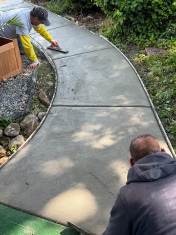Two people smooth wet concrete on a curved outdoor pathway surrounded by plants and stones.