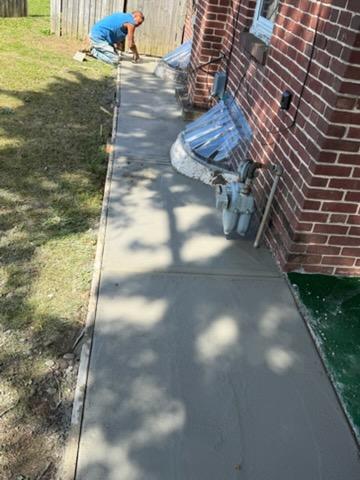 A person smooths wet concrete on a newly poured sidewalk beside a brick house.