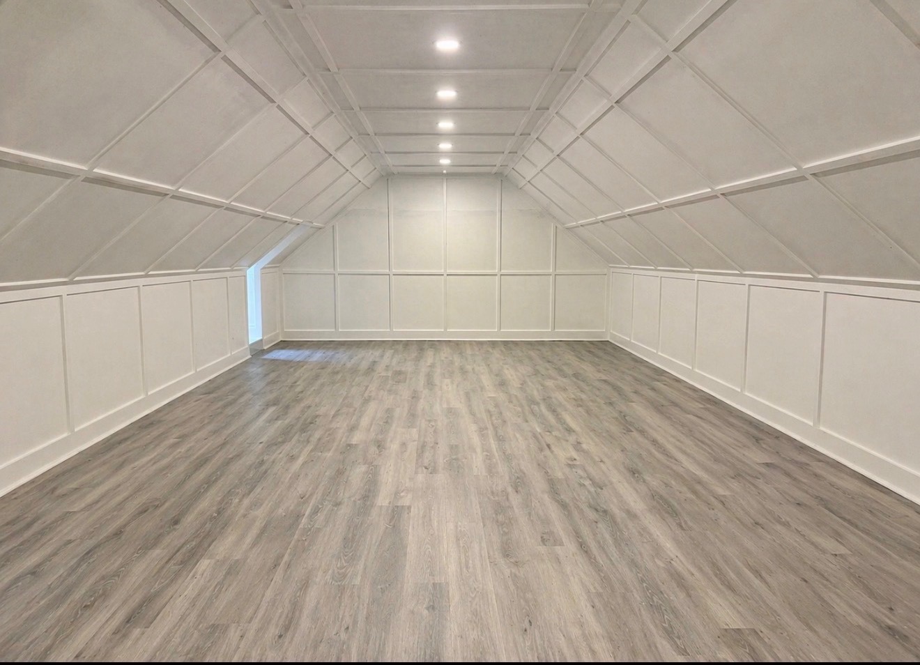 Bright, empty attic room with white paneled walls, recessed lighting, and wood-look flooring.