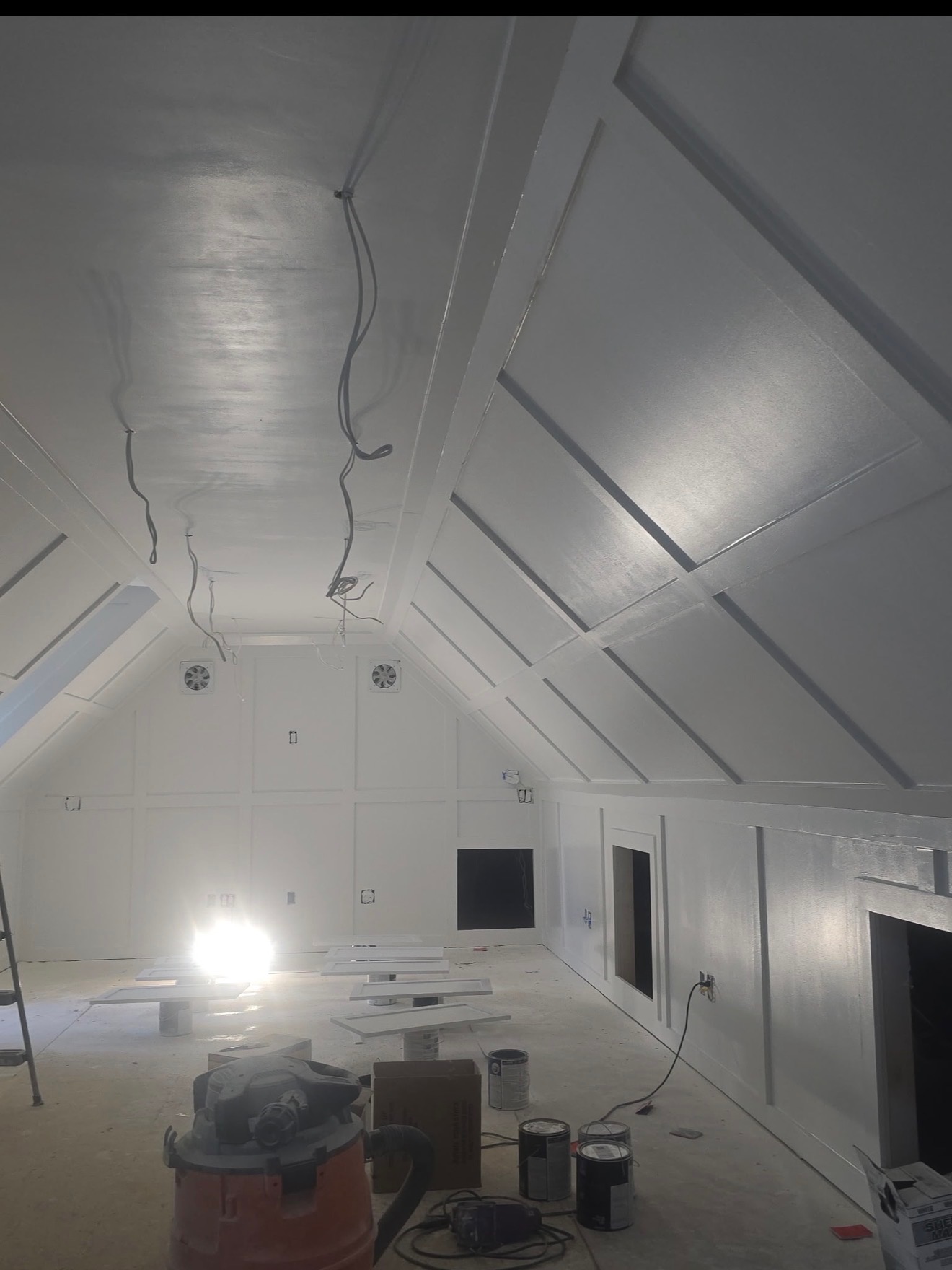 An unfinished attic with white paneled walls, exposed wiring, paint cans, and construction tools on the floor.