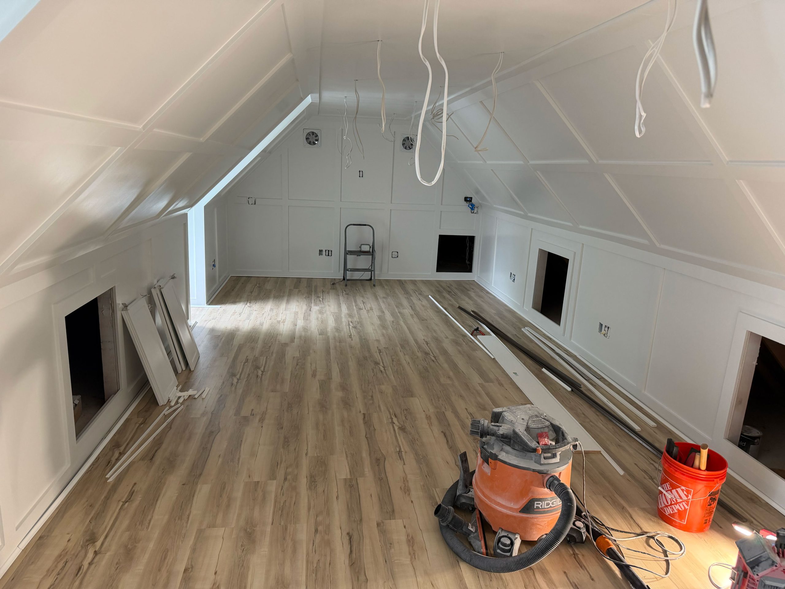 Unfinished attic room with wood flooring, white walls, tools, wiring, and construction materials scattered around.