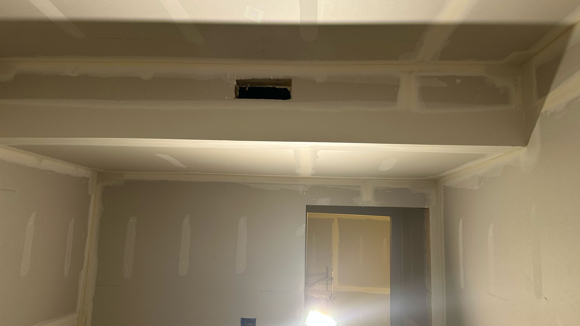 Unfinished room with drywall seams and joint compound, a ceiling vent opening, and light shining from below.