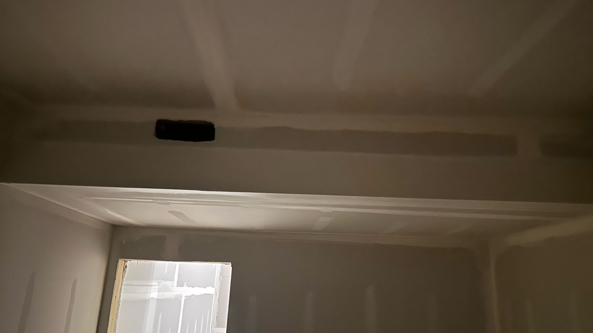 A dimly lit room with unfinished drywall and a small, rectangular vent near the ceiling.