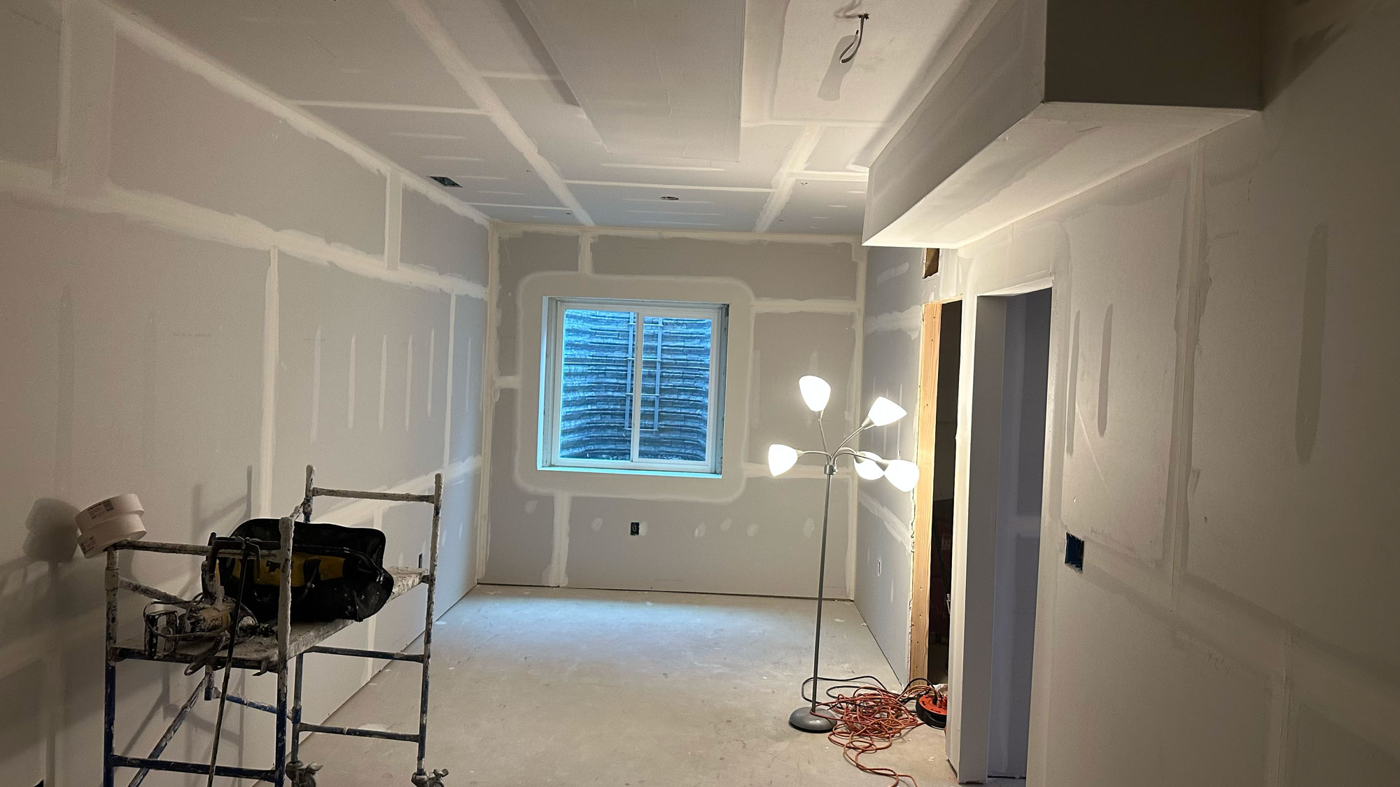 Unfinished room with drywall, a window, construction tools, and a lit floor lamp in the corner.
